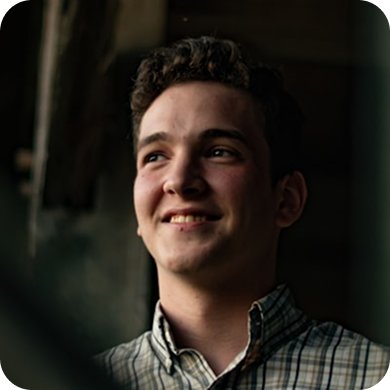 Young man with curly hair wearing a checkered shirt smiling while looking to his left.
