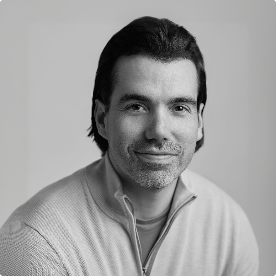 Smiling Justin Ernest with short dark hair wearing a light purple button-up shirt sitting with hands clasped.