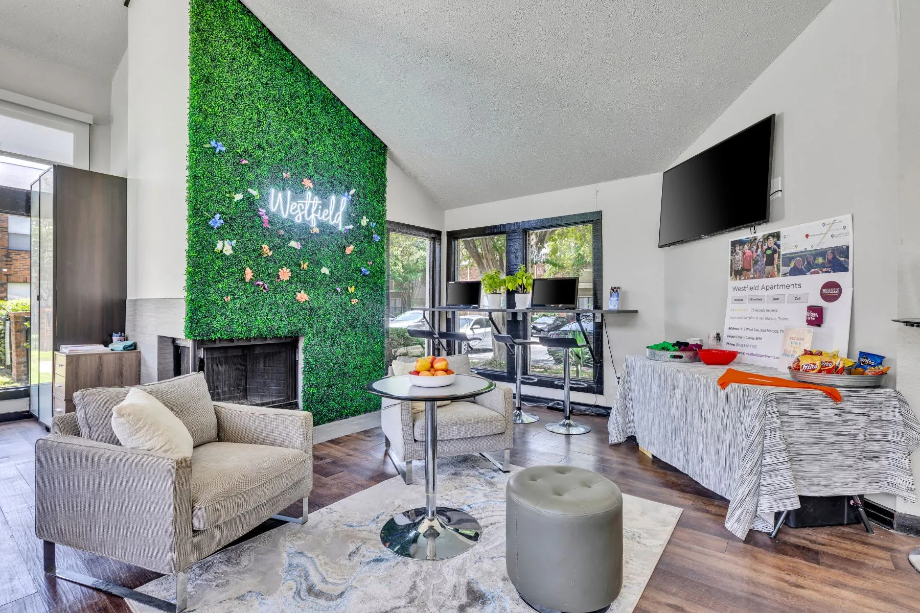 Lounge sitting area with grass wall, chairs, tv, table with fruit and desks with computers