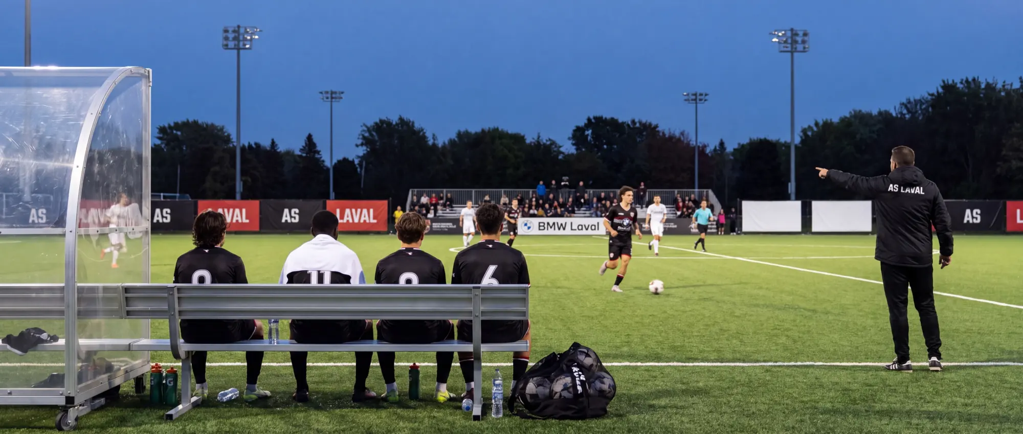 Joueurs de l’AS Laval sur le banc pendant un match semi-pro, devant les panneaux des partenaires du club