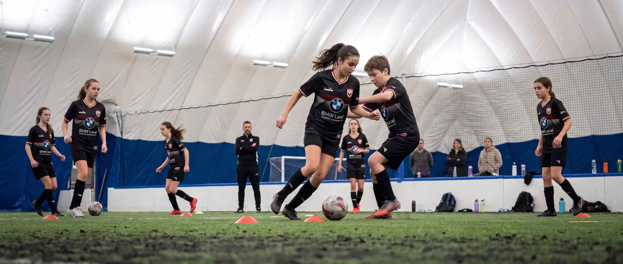 Joueuses et joueurs de l’AS Laval à l’entraînement sous le dôme, une joueuse dribble un adversaire entre les cônes