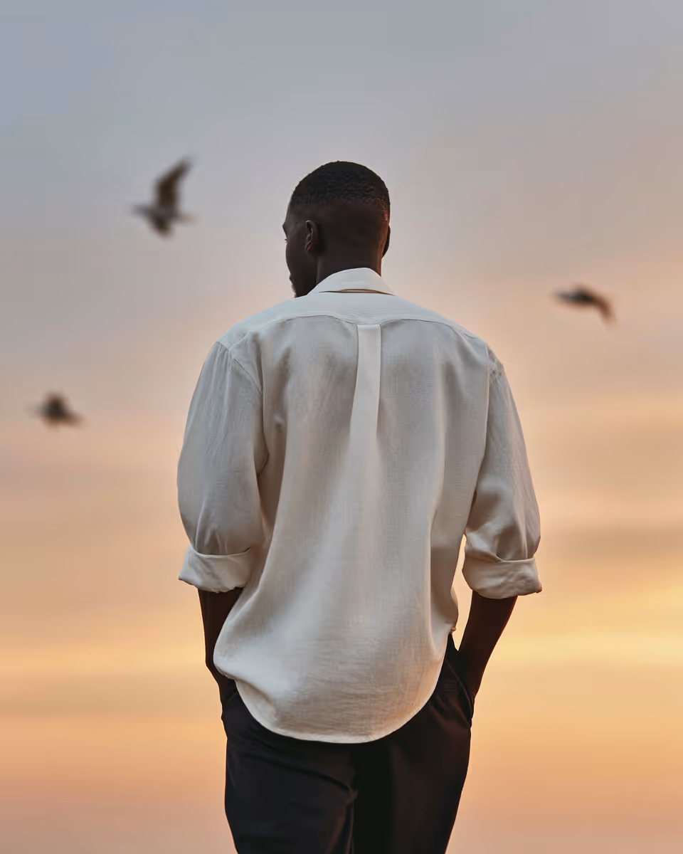 Man wearing a white shirt with rolled-up sleeves standing with hands in pockets, facing a sunset with birds flying.