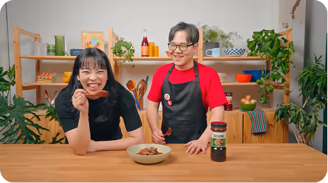 Irene holding a piece of cooked meat next to James in a red shirt and black apron, standing behind a wooden table with a bowl of food and a jar of seasoning.