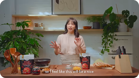 Irene standing at a kitchen counter with cooking ingredients and utensils, explaining a recipe with plants and shelves in the background.