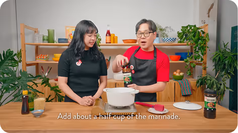 Irene and James cooking in a kitchen, with James holding a bottle of marinade over a pot.