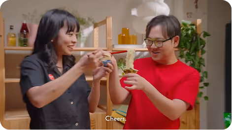 James and Irene smiling, holding chopsticks and a cup of noodles in a kitchen setting, with the subtitle 'Cheers.'