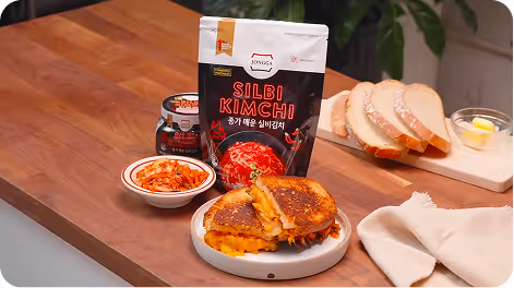 Grilled cheese sandwich cut in half on a plate with a packet of Silbi Kimchi, kimchi in a bowl, jar, sliced bread, and a small dish of butter on a wooden countertop.