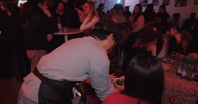James wearing a gray sweatshirt and black apron hands a bowl of food to a seated customer in a dimly lit, crowded restaurant.
