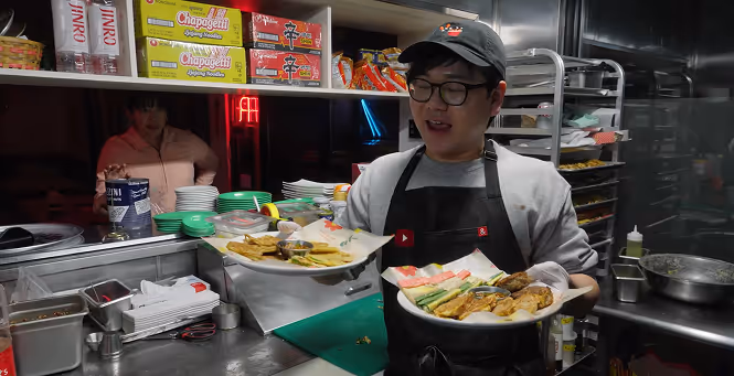 James wearing glasses and a black apron serving plates of assorted Korean food in a kitchen.