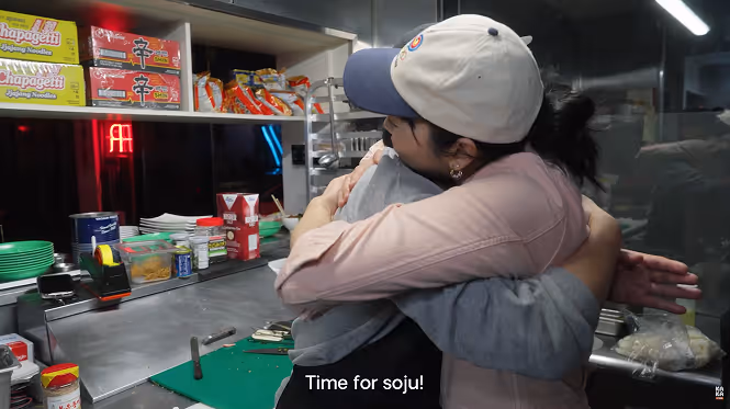 James and Irene hugging inside a kitchen with shelves of instant noodles and kitchen supplies in the background.