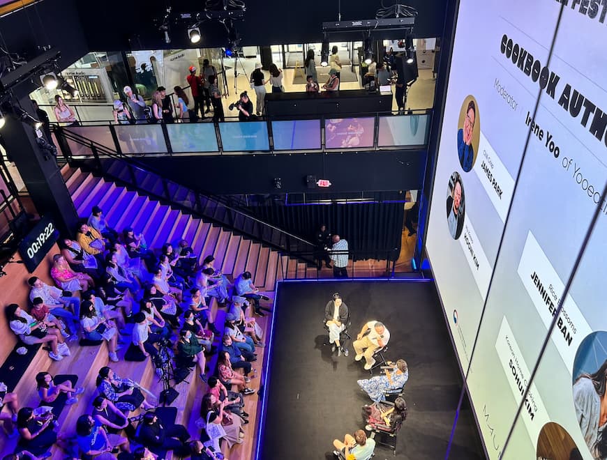 An overhead view of a panel discussion on a stage with six speakers seated, an audience seated on wooden steps, and a large screen displaying cookbook author names and photos in a modern event space.