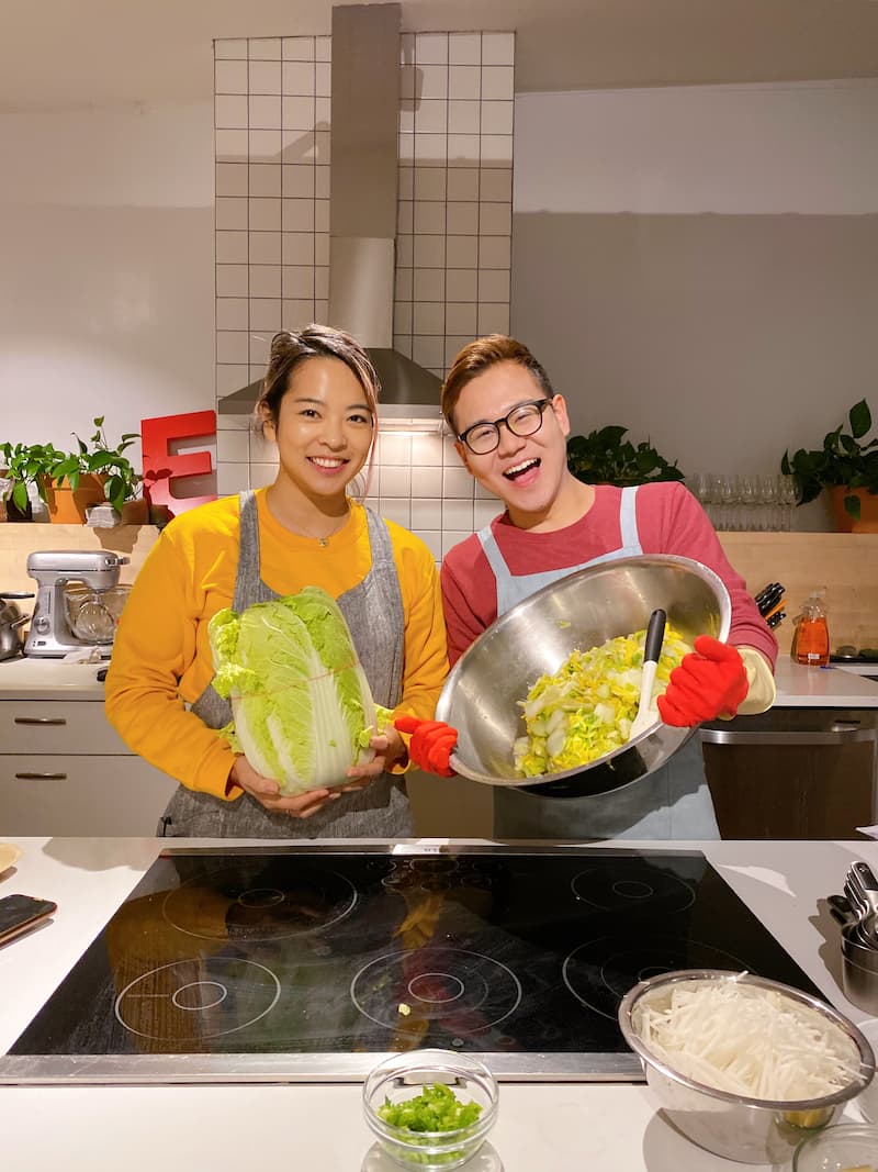 Irene and James in aprons standing in a kitchen, Irene holding a whole napa cabbage, James holding a large bowl of chopped cabbage and wearing red gloves.