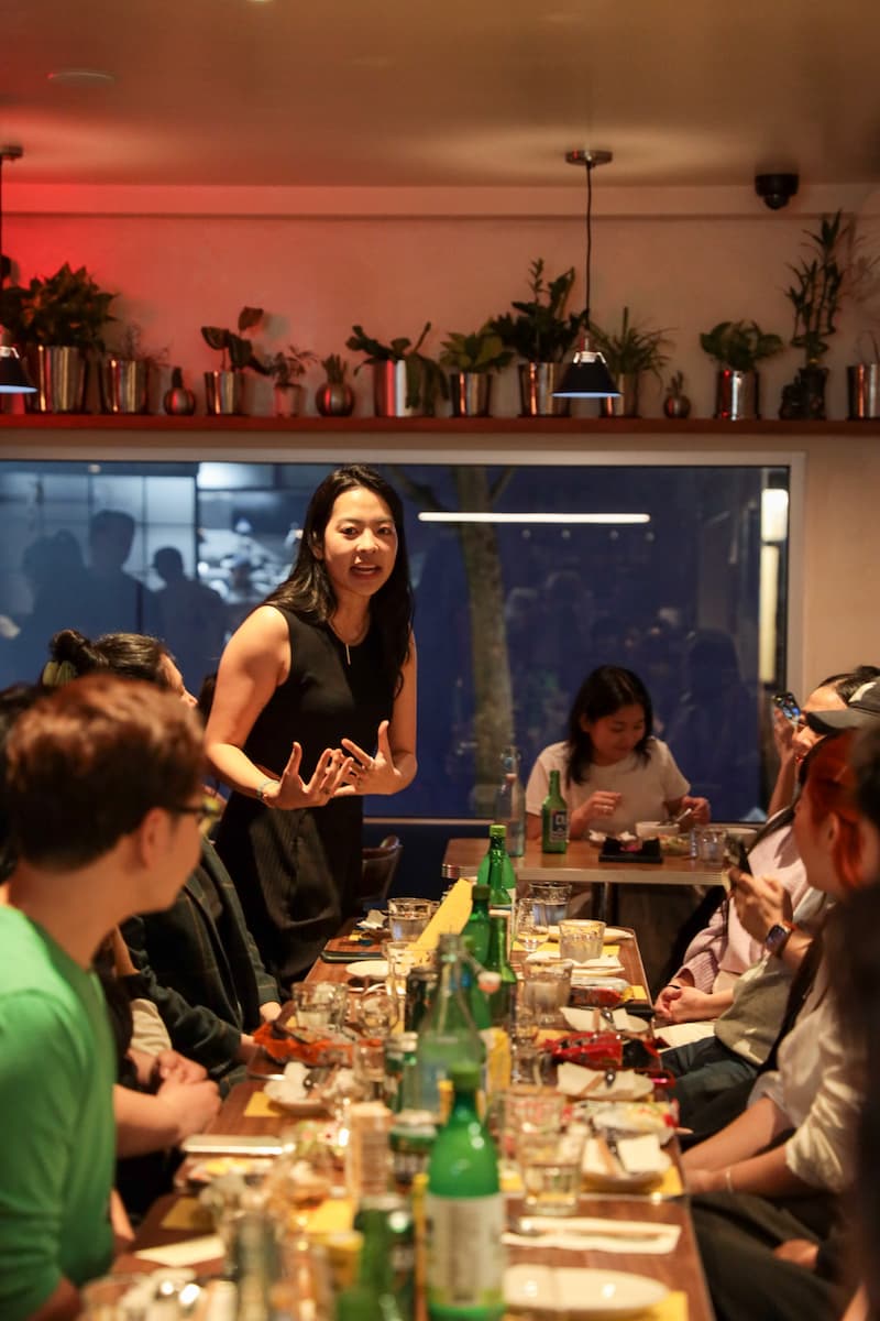 Irene standing and speaking animatedly to a group seated around a long table covered with drinks and dishes in a warmly lit room.