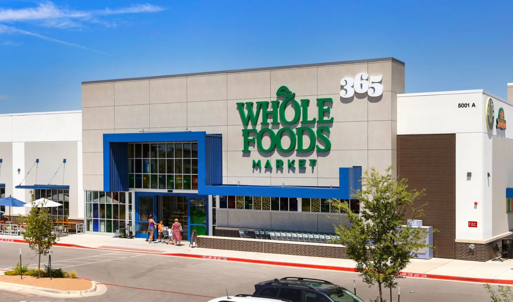Exterior of a Whole Foods Market 365 store with entrance, blue accents, and several people walking nearby.