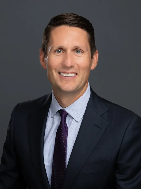 Smiling man in a dark suit, light blue shirt, and dark purple tie against a gray background.