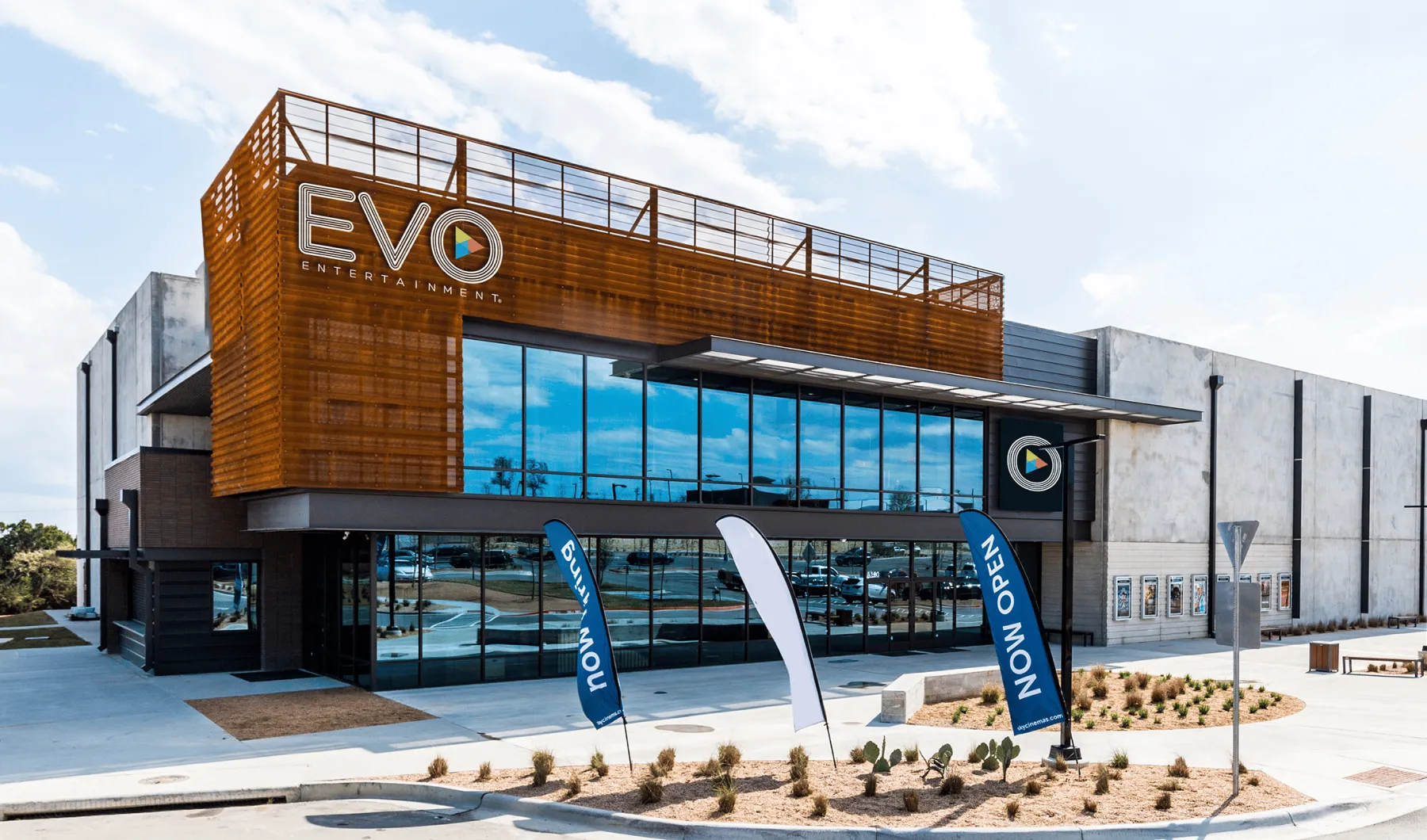 Modern commercial building with large reflective glass windows and brown metal paneling, featuring EVO Entertainment signage and 'Now Open' blue banners in front.