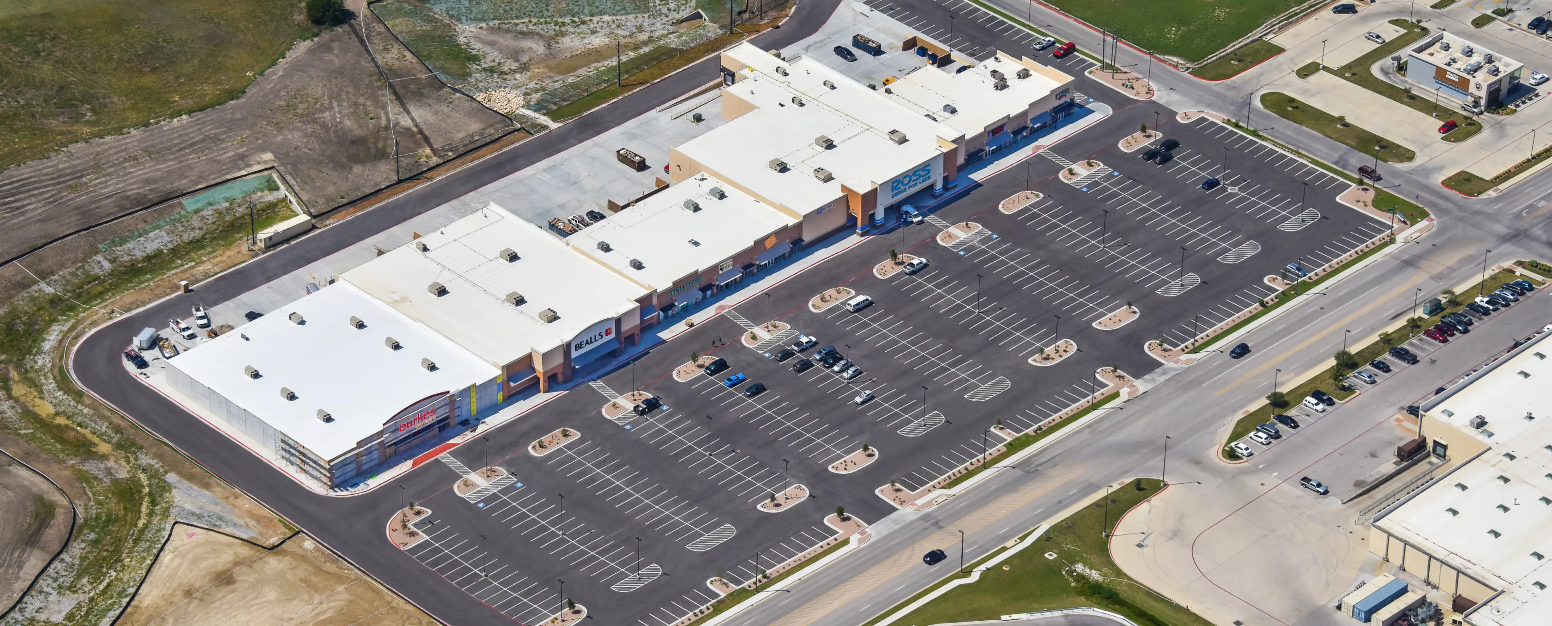 Aerial view of a commercial area including two large retail buildings under construction along a busy highway, surrounded by parking lots, smaller stores, and residential neighborhoods.