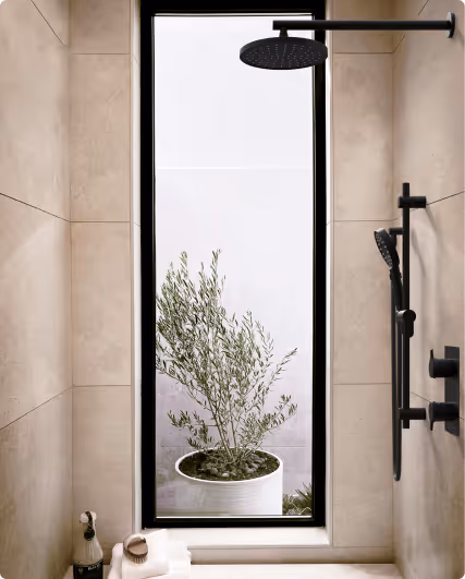 Modern shower area with beige tiles, black rainfall showerhead, handheld shower, and a tall window showing a potted plant outside.