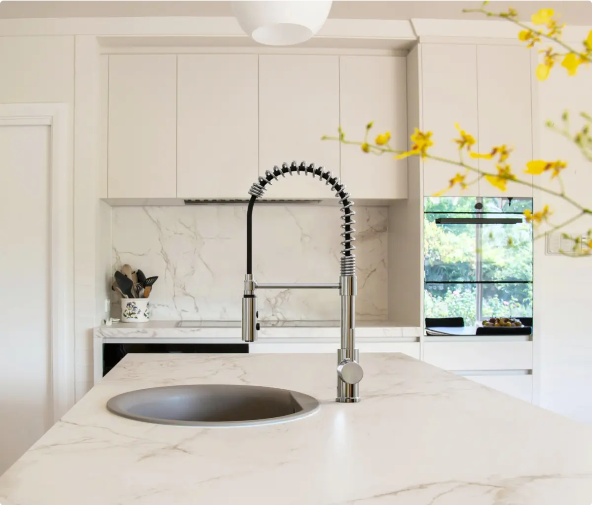 Modern kitchen with a sleek silver spring-style faucet over a round sink set in a white marble countertop and yellow flowers blurred in the foreground.