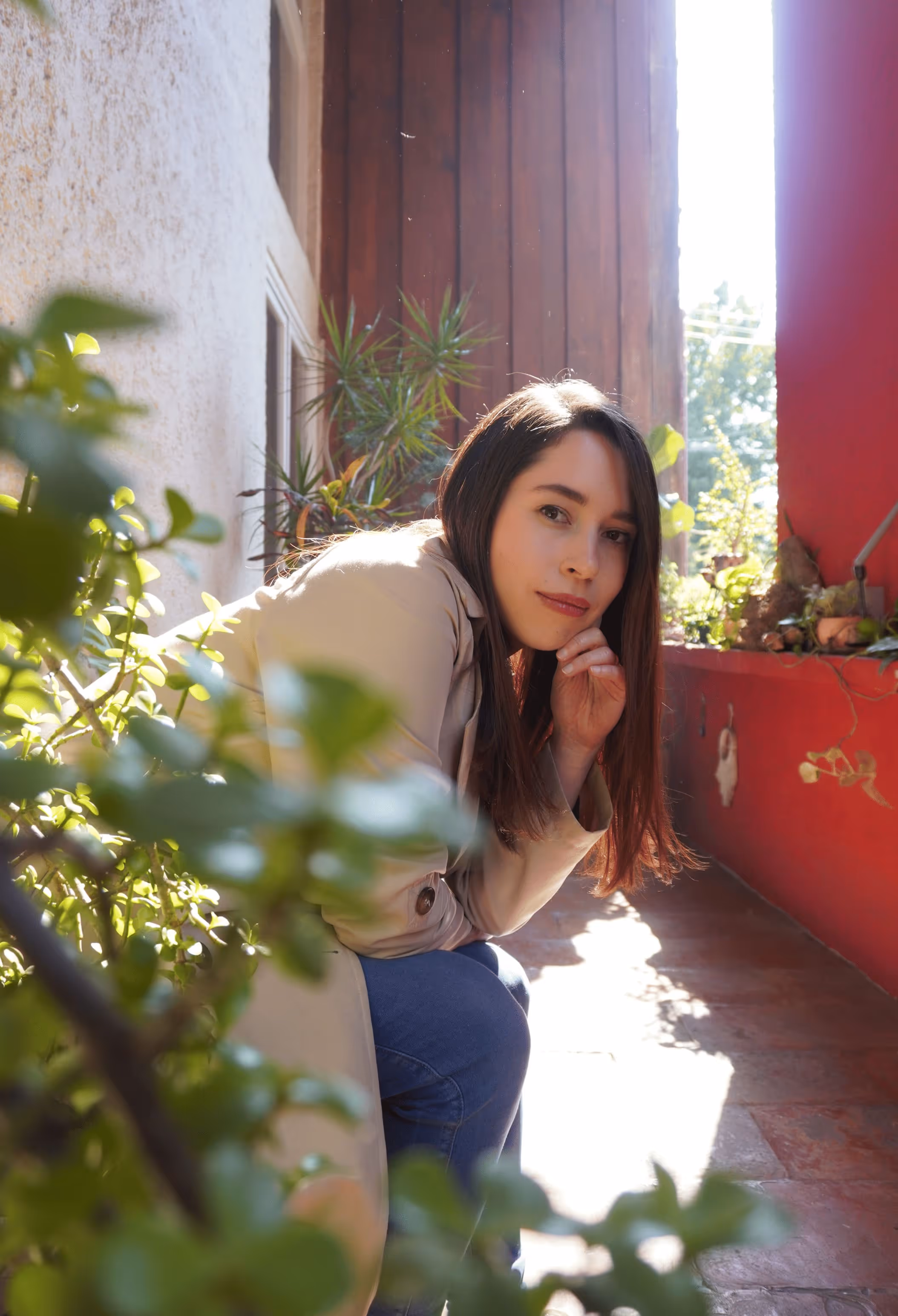 Young woman with long brown hair wearing a beige coat and blue jeans, crouching in a sunlit corridor with plants around.