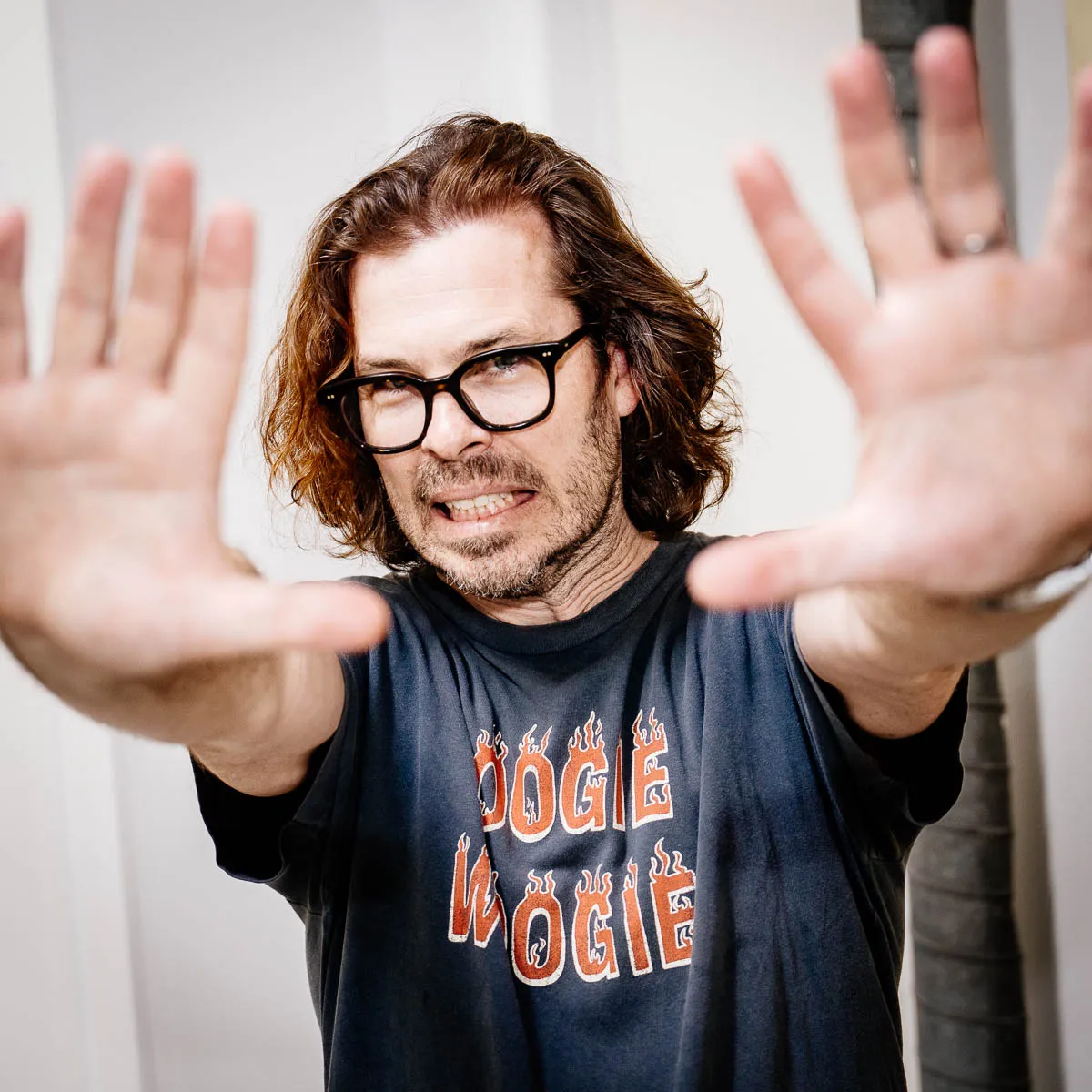 Man with shoulder-length hair and glasses reaching hands toward the camera wearing a dark t-shirt with 'BOOGIE WOOGIE' text.