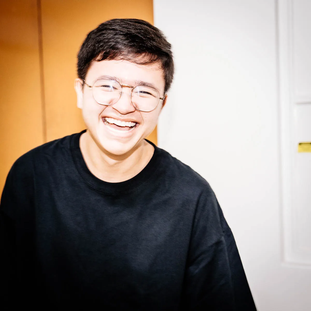 Young person with short dark hair and glasses laughing while wearing a black shirt.