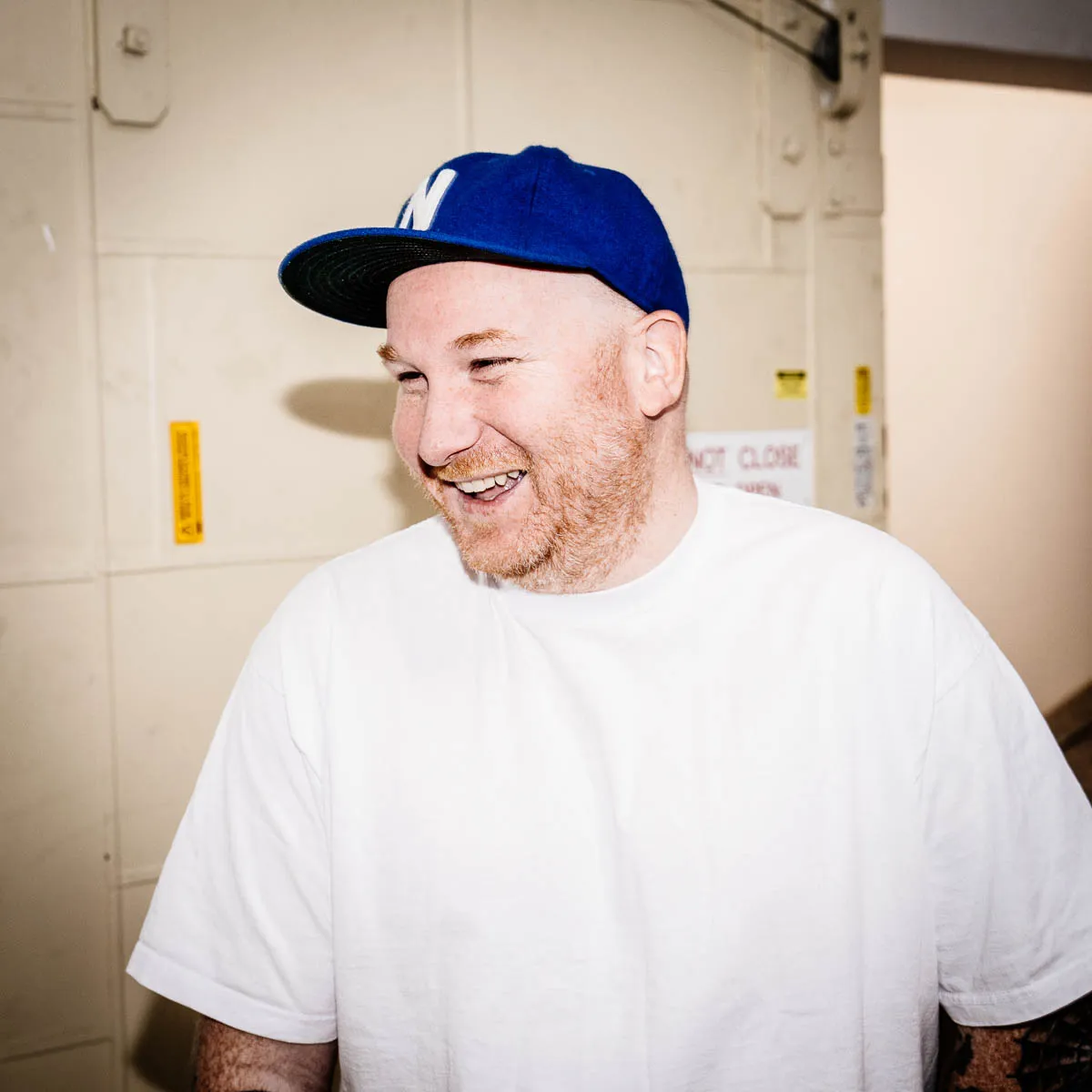 Smiling man with a red beard wearing a blue cap and white t-shirt standing indoors.