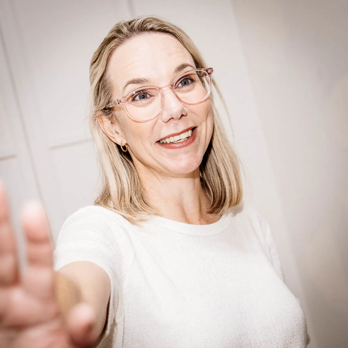 Smiling woman with blonde hair and pink glasses reaching her hand forward.