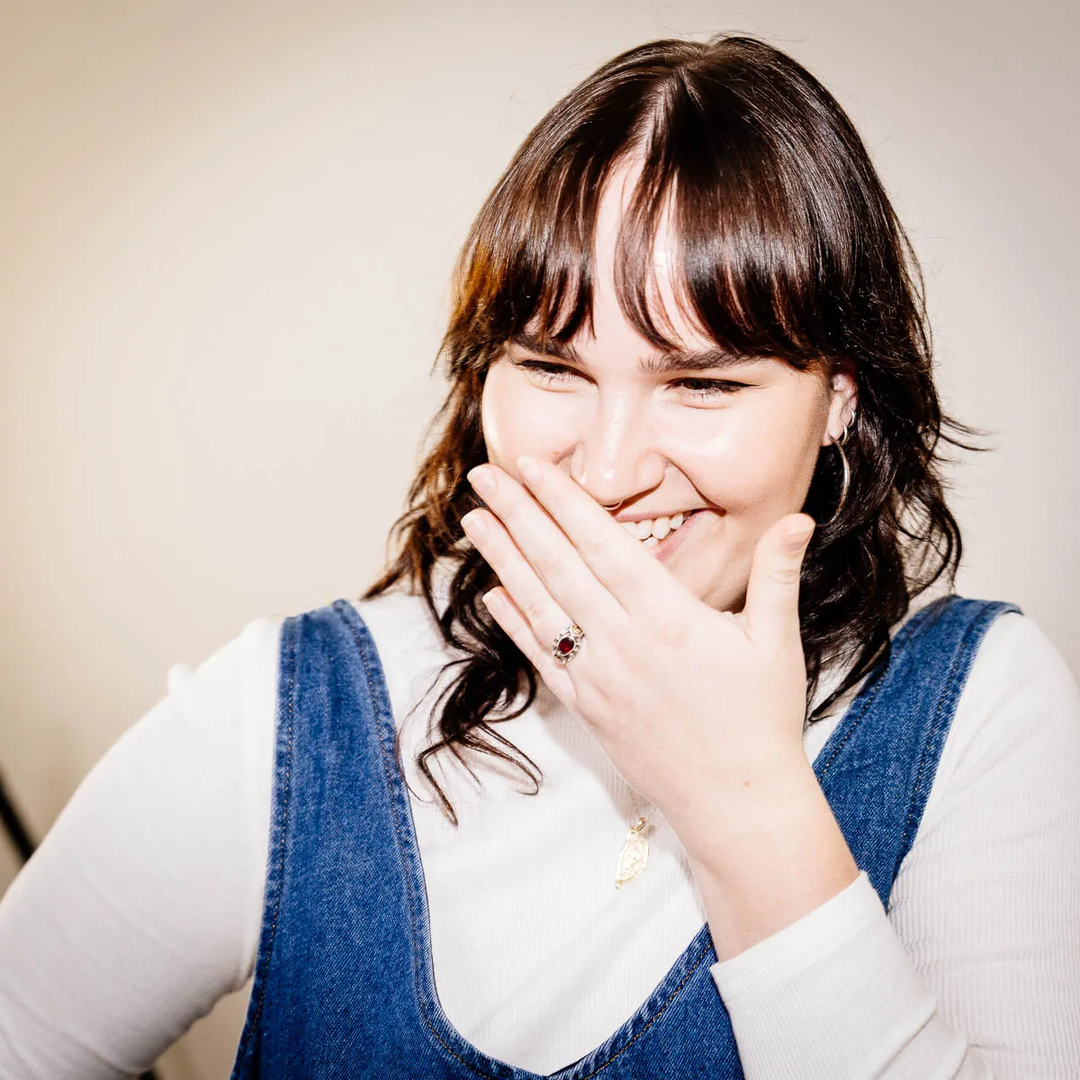 Smiling woman with dark hair covering her mouth with her hand while wearing a white long-sleeve shirt and blue denim dress.