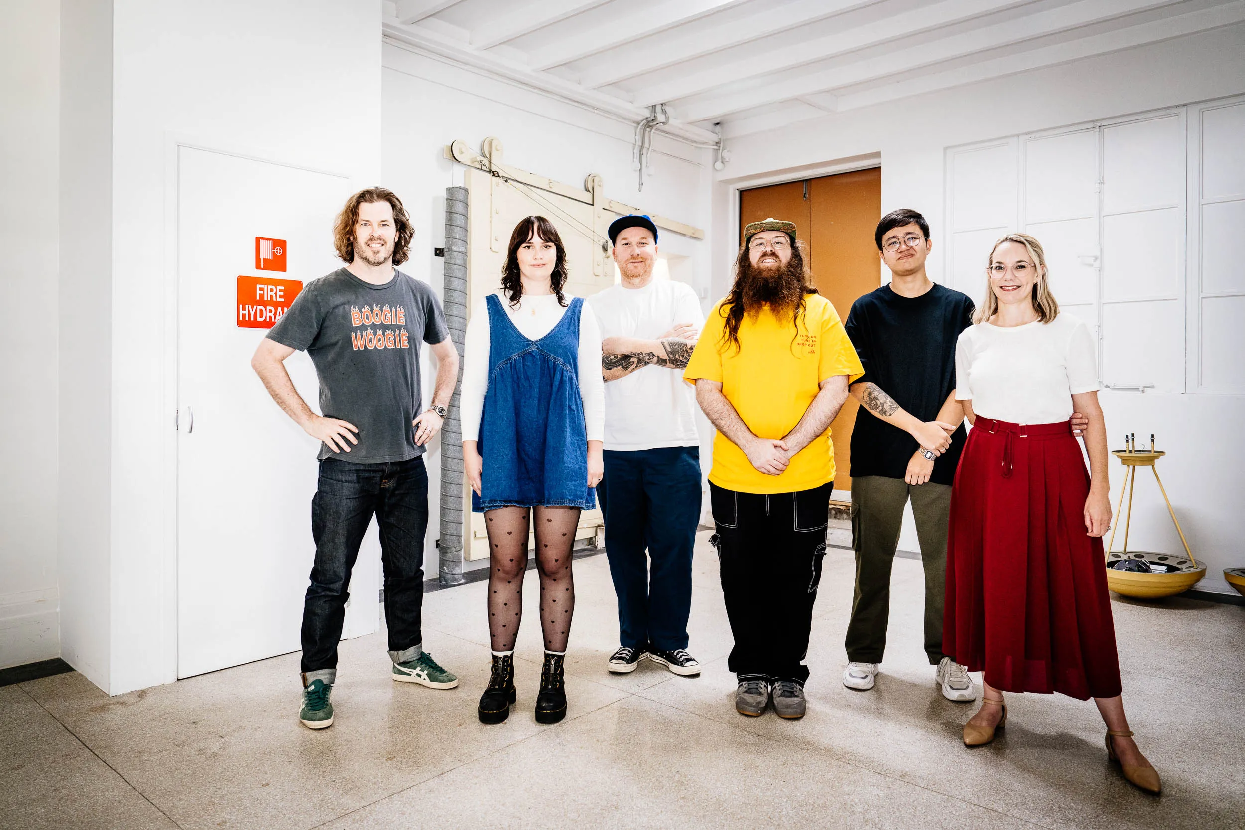 Group of six people standing indoors against a white wall, casually dressed, smiling at the camera.
