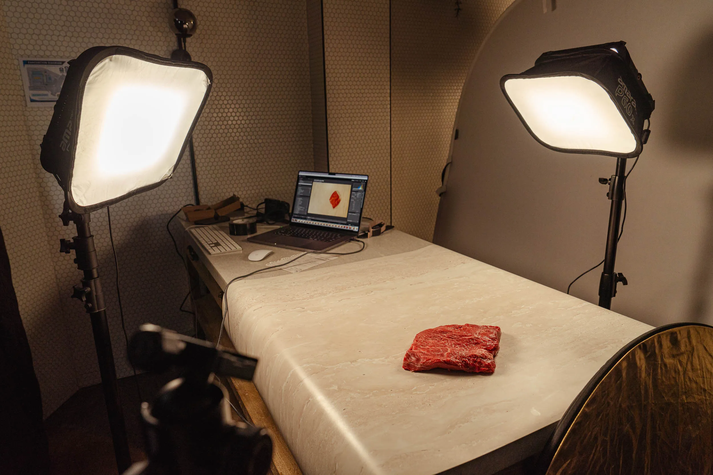 Photography setup with two softbox lights illuminating a piece of raw meat on a table, with a laptop displaying the meat in editing software.