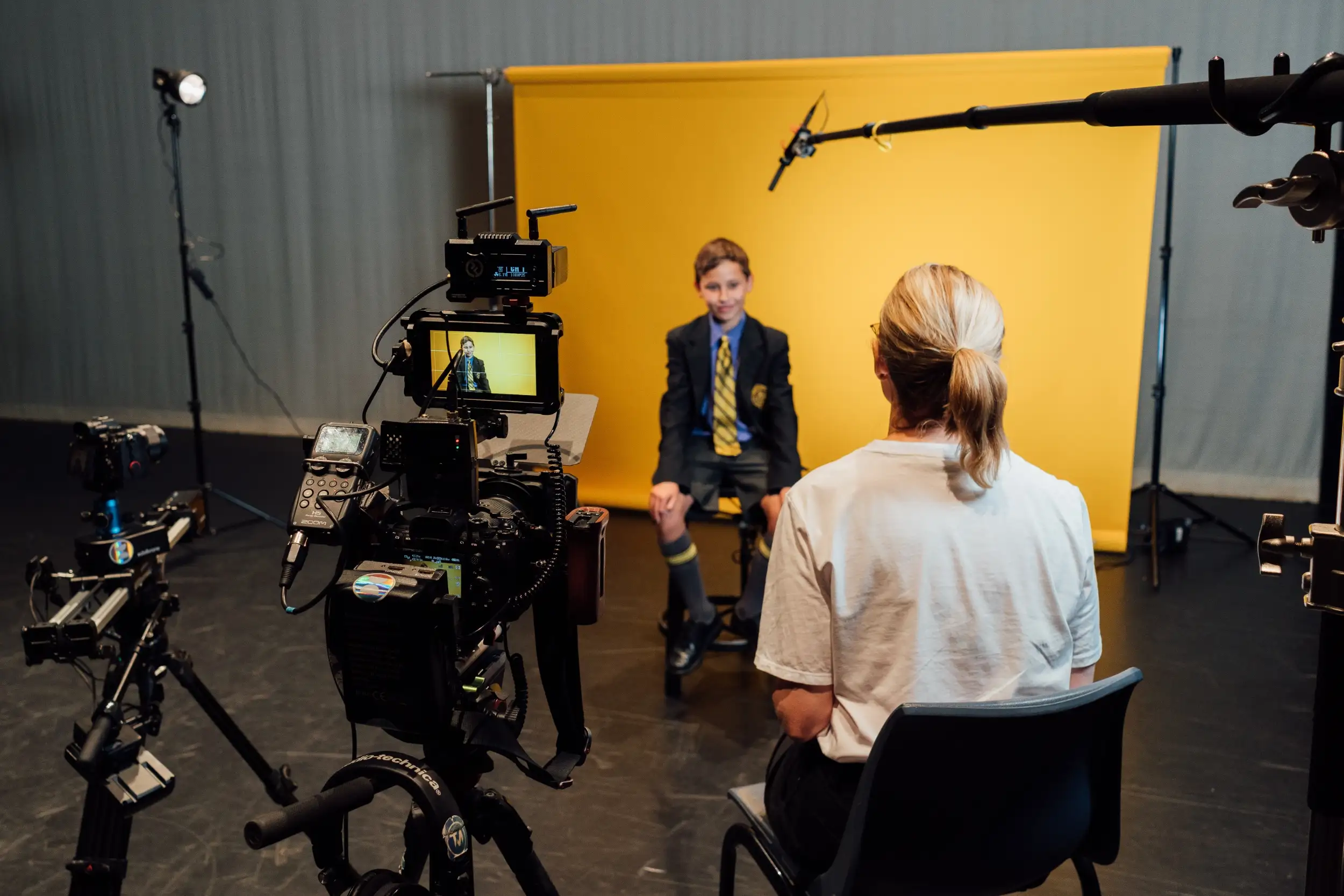 Boy in a school uniform being filmed against a yellow backdrop by a camera crew with a person interviewing him.