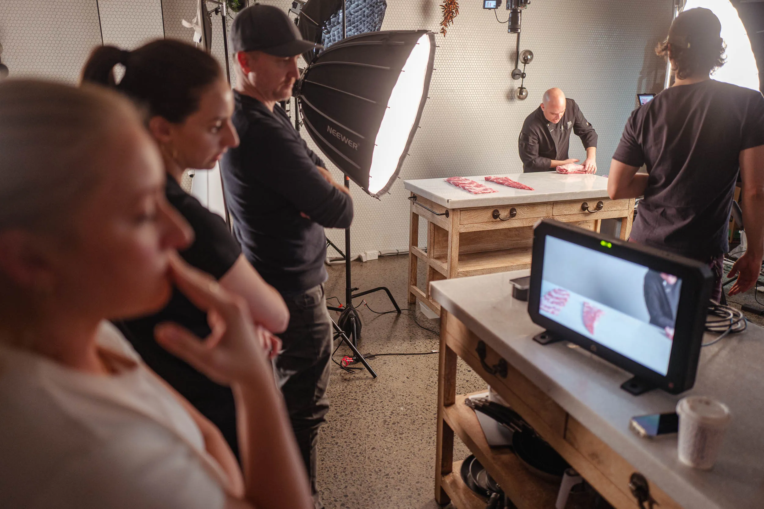 A chef demonstrating meat preparation on a table while a film crew with lighting and camera equipment films and watches.