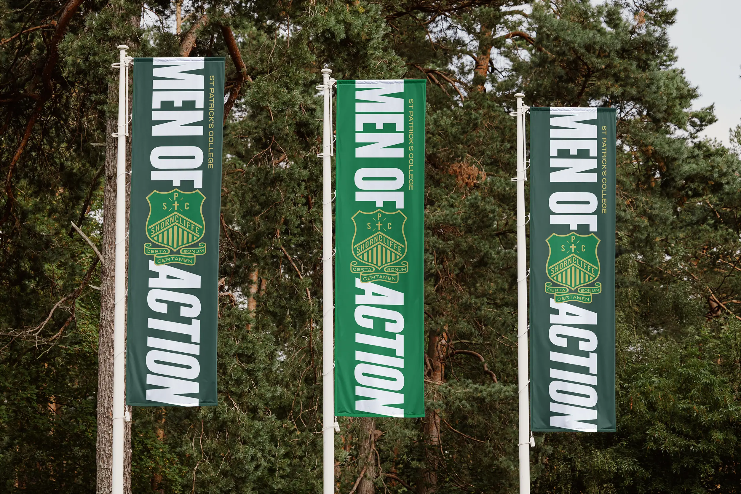 Three vertical green banners with white text reading 'MEN OF ACTION' and the St Patrick's College crest hanging from flagpoles among trees.