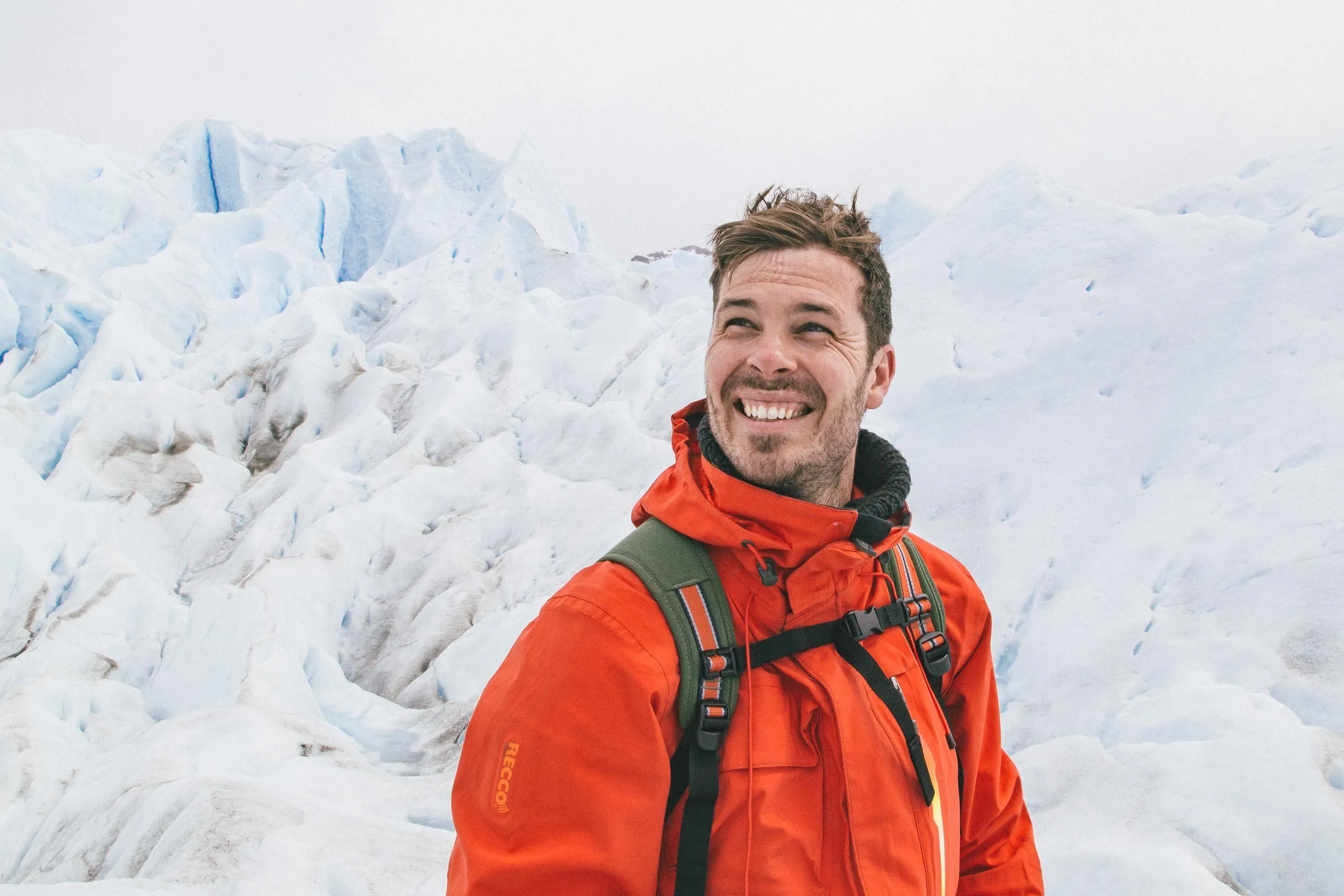 Smiling man wearing an orange jacket and green backpack standing in front of a glacier.