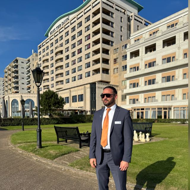 Portrait of a smiling man with dark hair, beard, and suit jacket against a dark background.