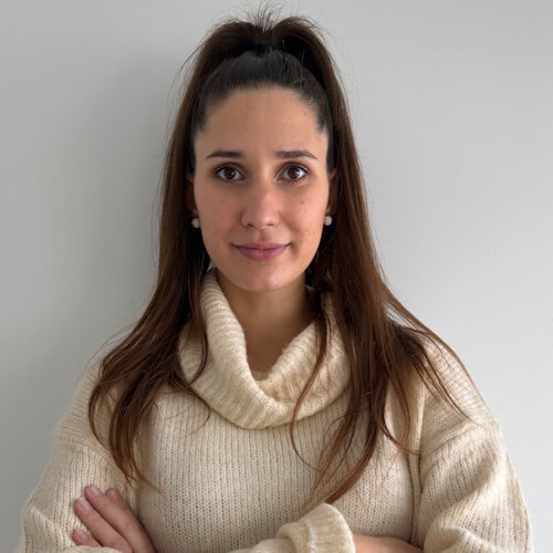 Ljubica - Woman with long brown hair in a high ponytail wearing a cream turtleneck sweater, standing against a plain light gray background with her arms crossed.