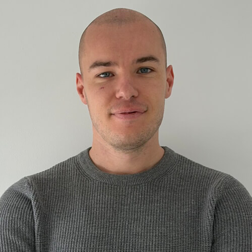 Marko -Smiling man with a shaved head wearing a grey knitted sweater, standing against a plain white wall.