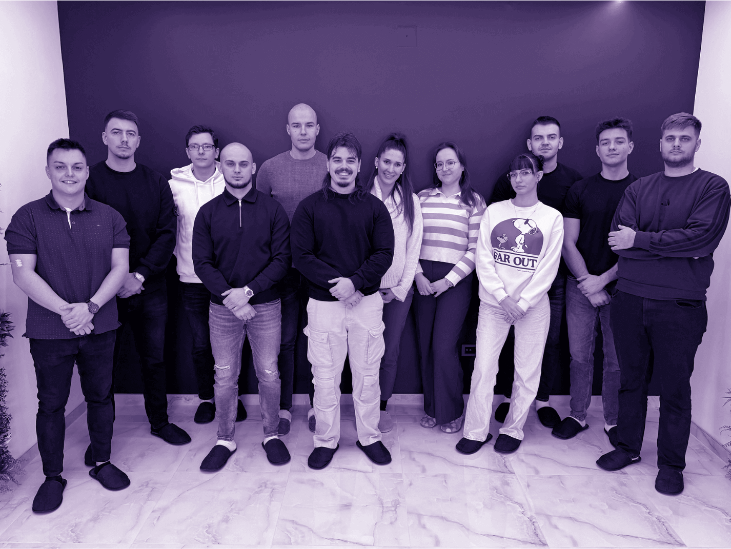 Group of thirteen people standing indoors against a dark purple wall, posing for a team photo on a marble floor.