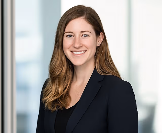 Smiling woman with long brown hair wearing a black blazer in a bright office setting.