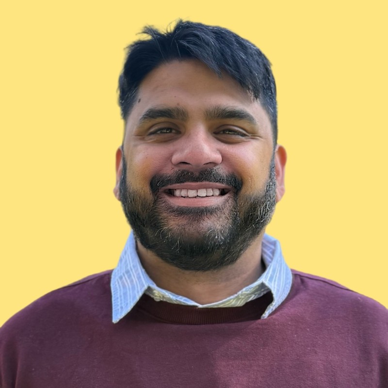Smiling man in a beige suit jacket and white shirt standing in a modern office with a desk, computer, and floor lamp in the background.