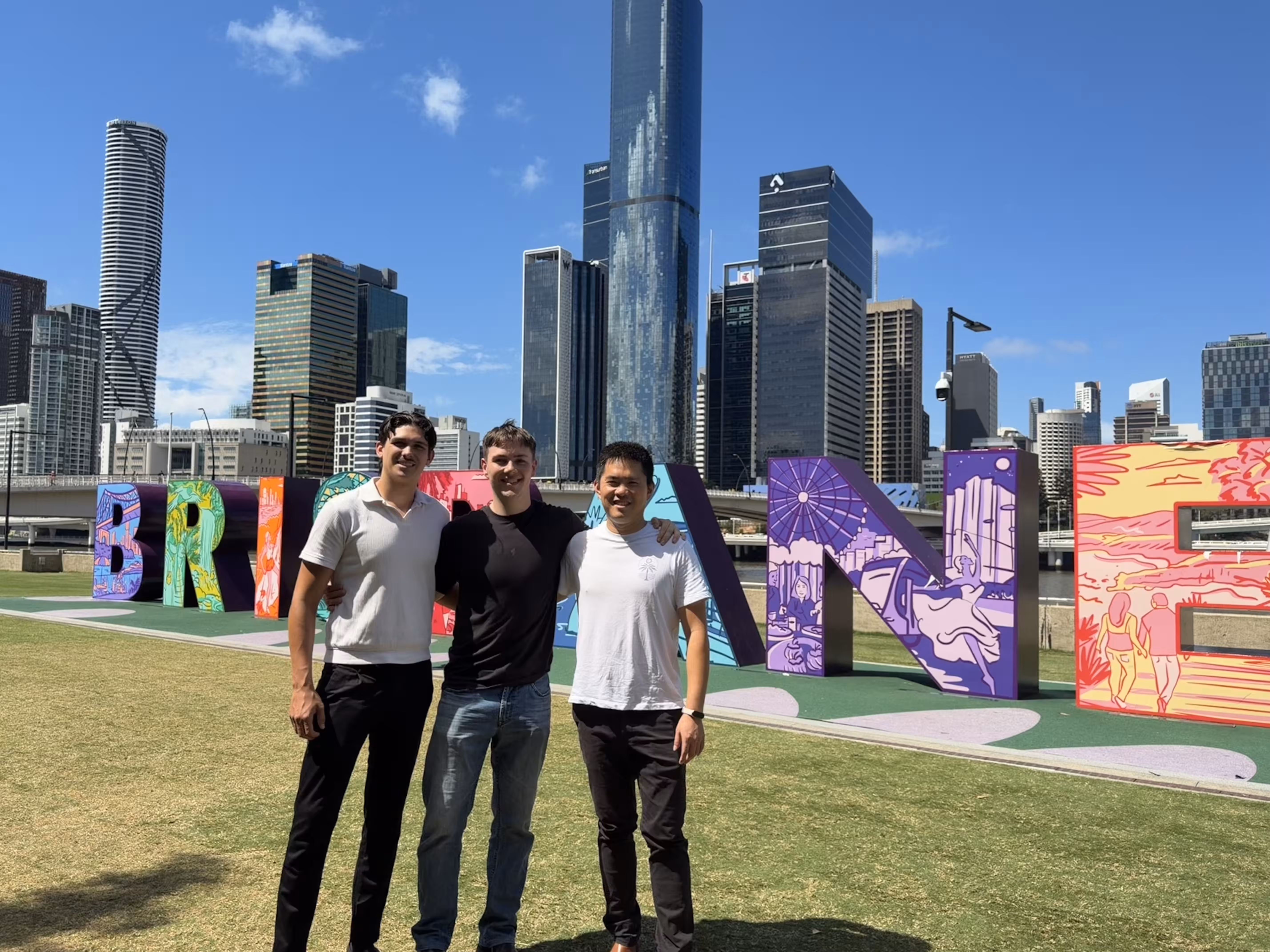 Three men standing on grass in front of colorful 'BRISBANE' letters with a city skyline and blue sky in the background.