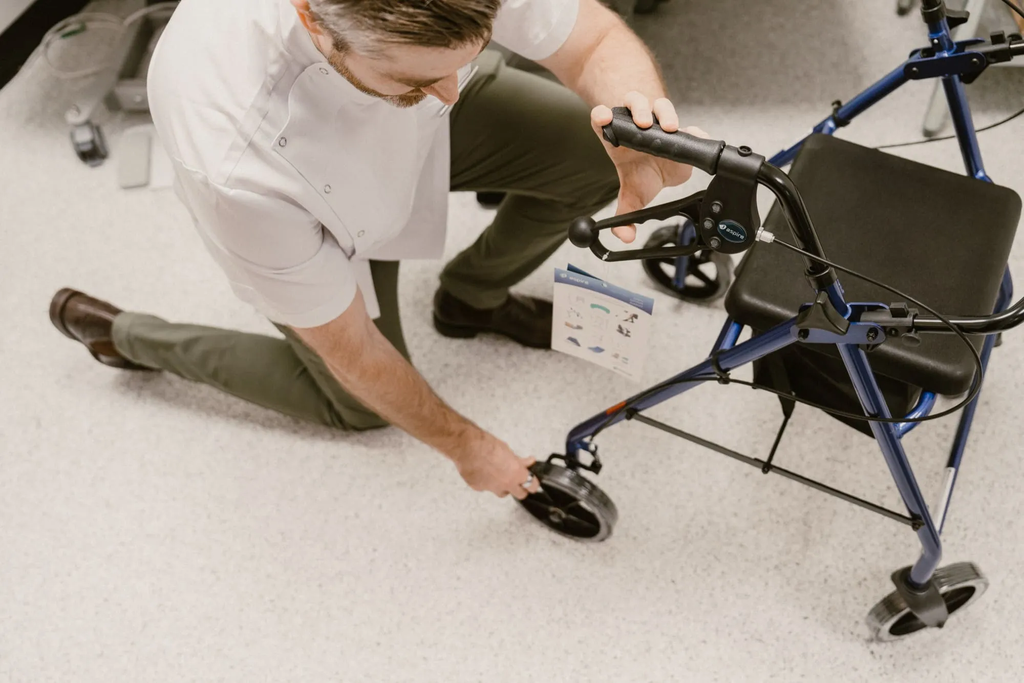 Man kneeling on the floor adjusting the wheel of a blue walker with a black seat in a clinical setting.