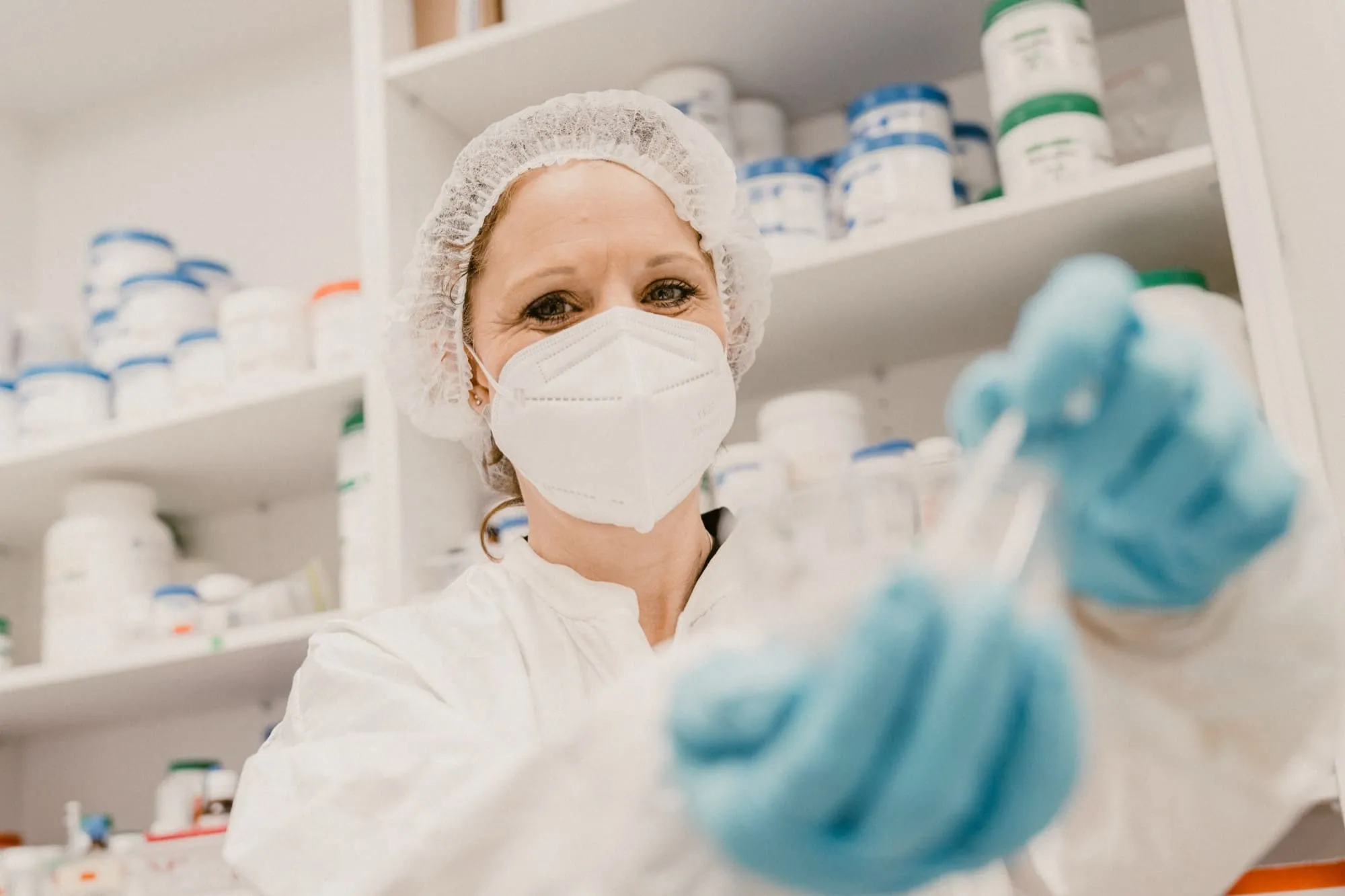 Female pharmaceutical worker in protective gear and mask handling medicine bottles in a pharmacy.