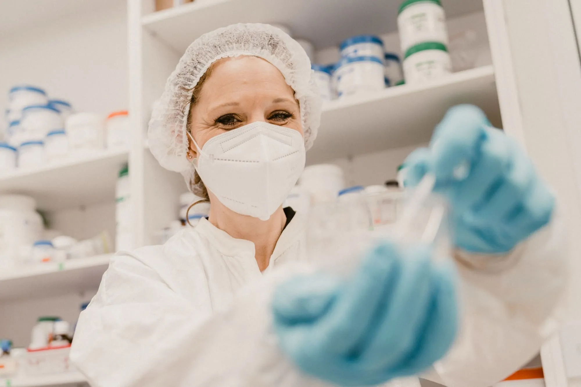 Pharmacist wearing protective gear including a hairnet, face mask, and gloves, preparing medication in a pharmacy.