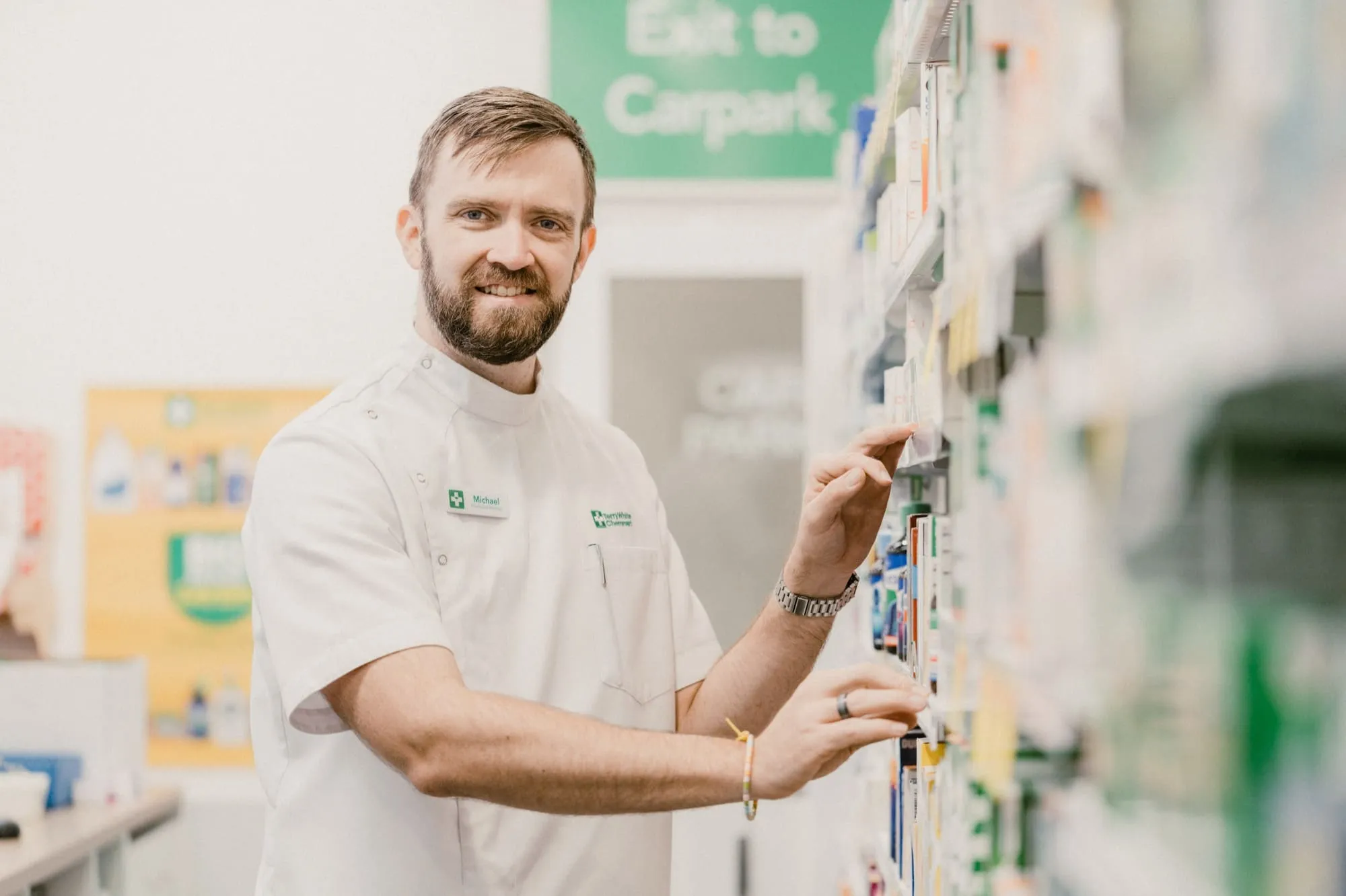 Pharmacist named Michael in white uniform organizing medication boxes on pharmacy shelves.