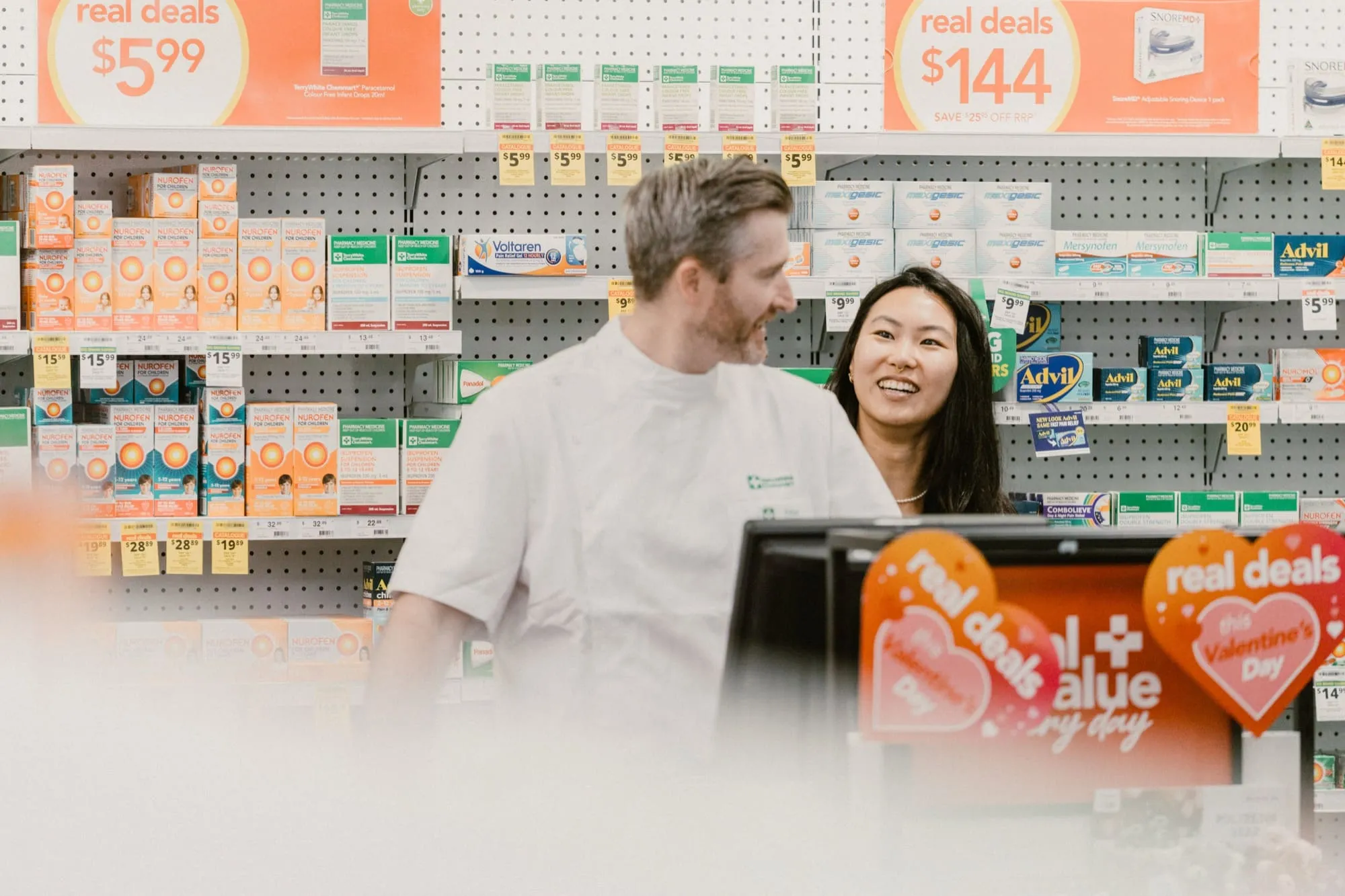 Pharmacy staff smiling and talking behind a counter with shelves of medicine and promotional signs in the background.