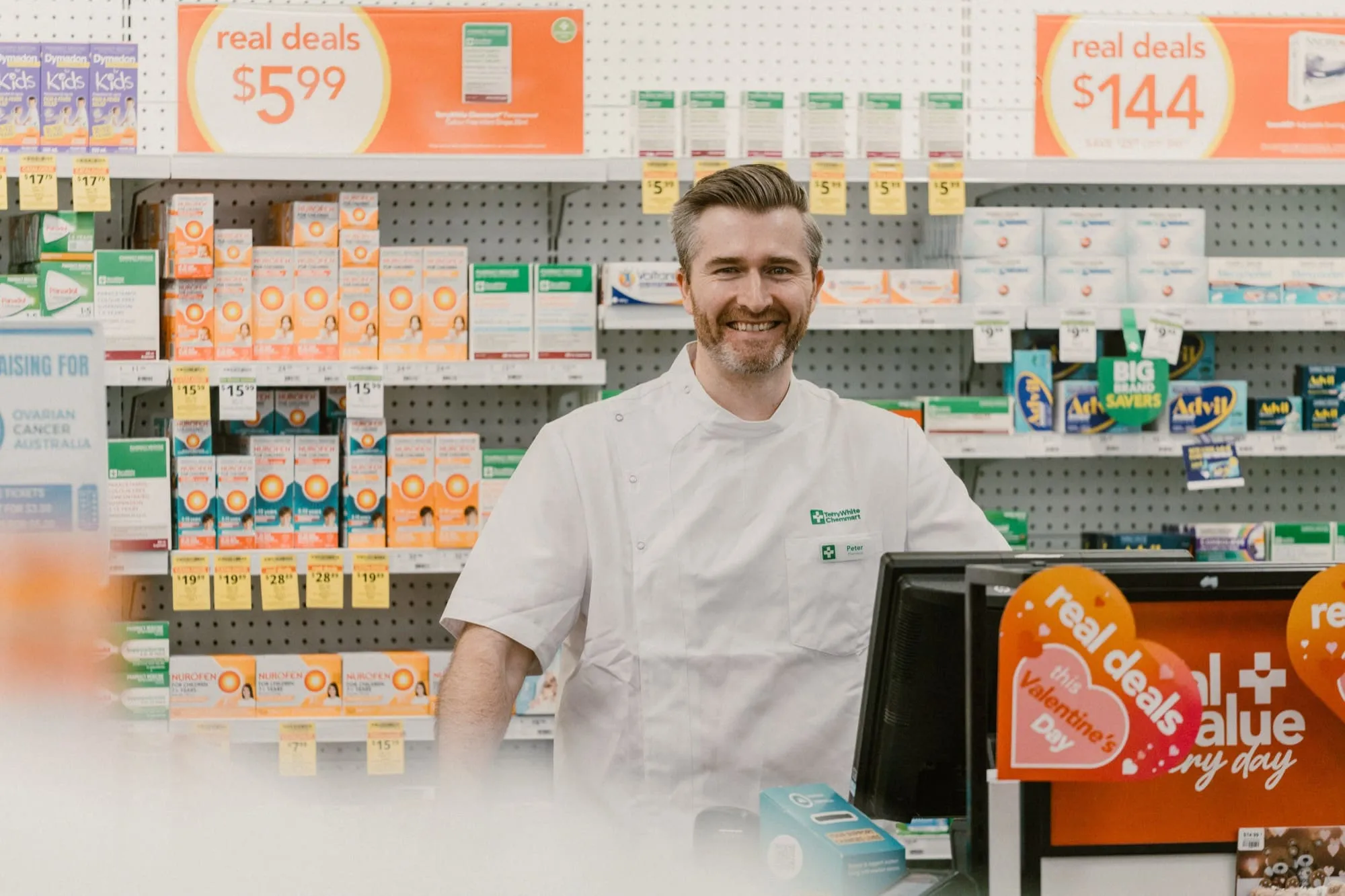 Smiling male pharmacist in white coat standing behind a pharmacy counter with shelves of health products and real deals signs in the background.