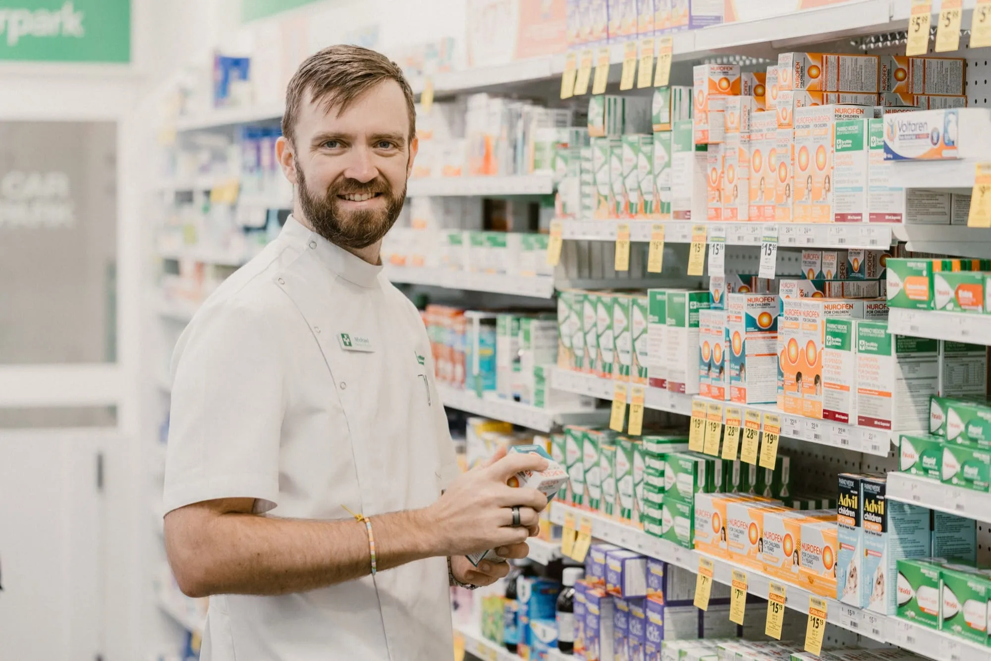 Smiling pharmacist in white coat standing by shelves stocked with over-the-counter medications.