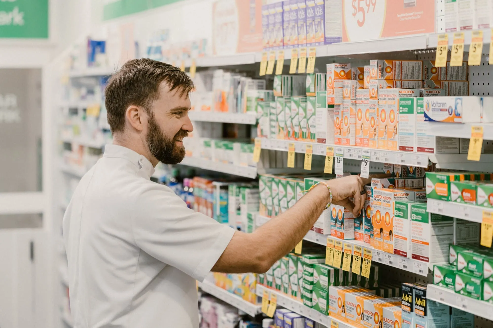 Smiling male pharmacist arranging medication boxes on a pharmacy shelf.